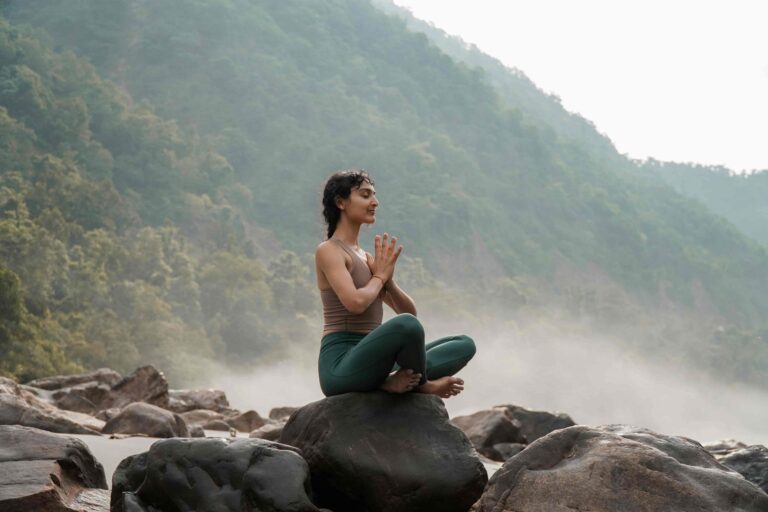 Woman practicing yoga in lotus pose on a rock in nature with mountains and mist in the background